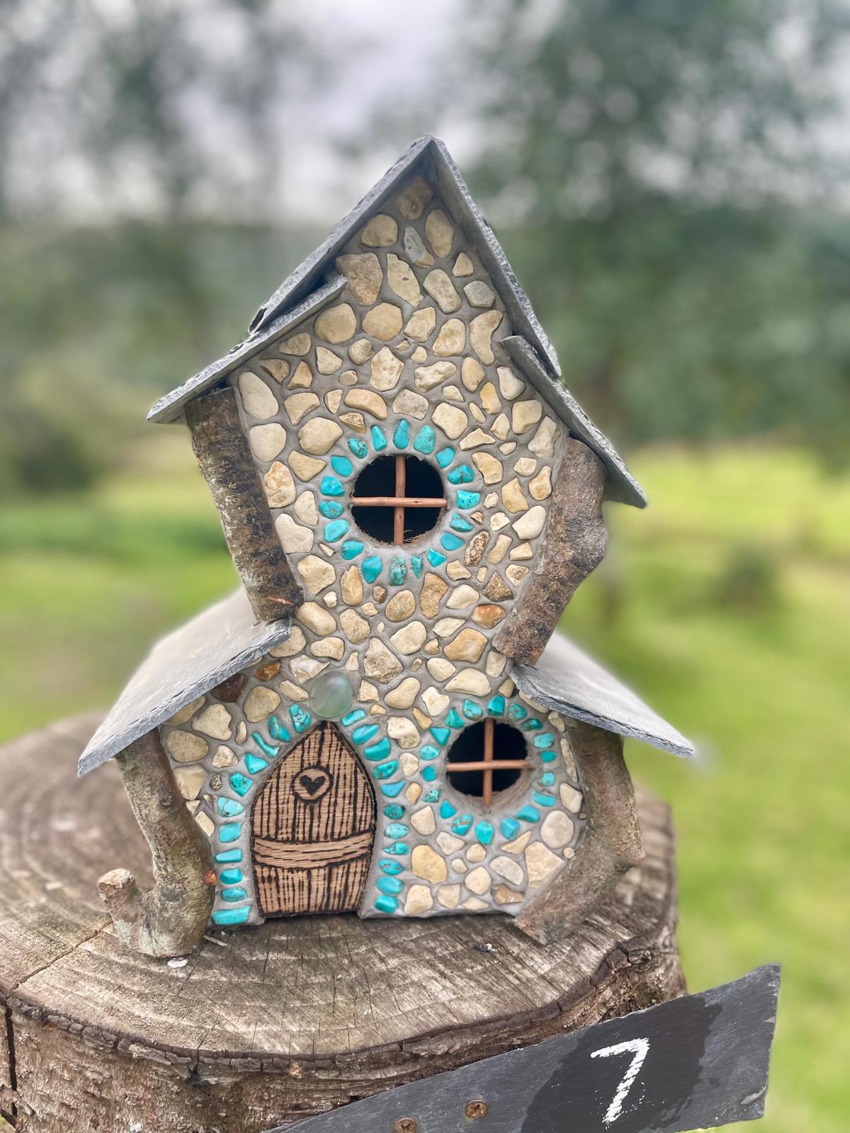 A miniature fairy house crafted from small beige and brown stones, featuring a dark gray slate roof, a brown wooden-textured door with a heart detail, and turquoise painted accents around the windows, displayed on a rustic tree stump with a blurred green outdoor background.