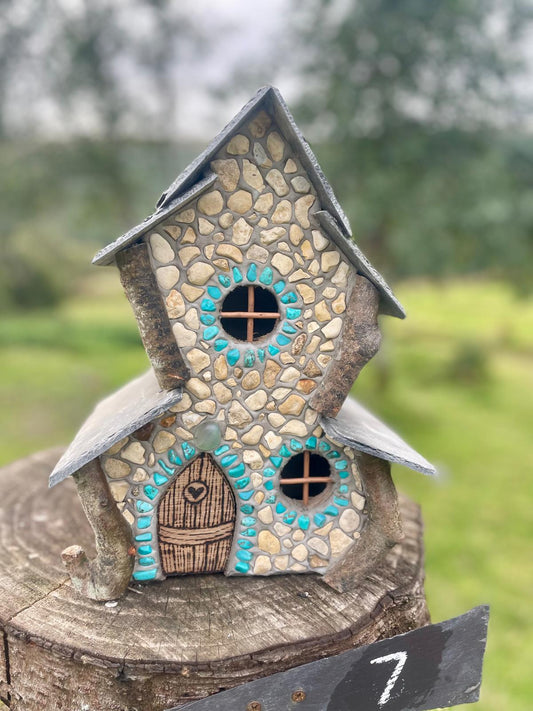A miniature fairy house crafted from small beige and brown stones, featuring a dark gray slate roof, a brown wooden-textured door with a heart detail, and turquoise painted accents around the windows, displayed on a rustic tree stump with a blurred green outdoor background.
